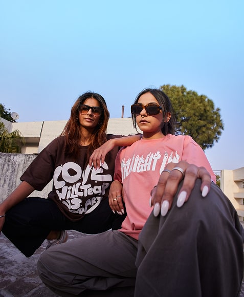 Two women in oversized crewneck tees, brown with white "LOVE" chest graphic and pink with centered flame text, low-angle shot
