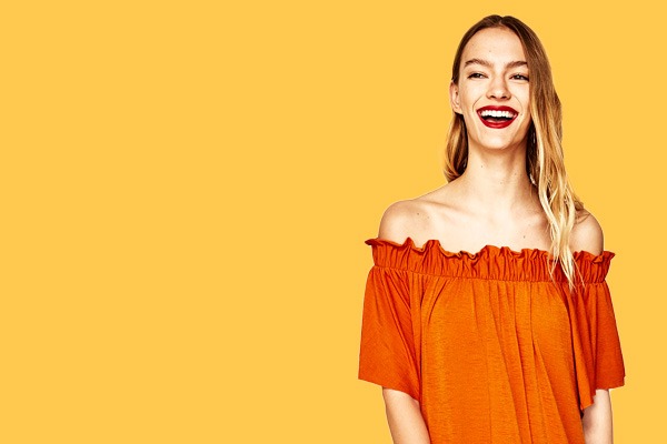 orange off-shoulder blouse loose fit with ruffled neckline front view model against yellow backdrop, center-right composition