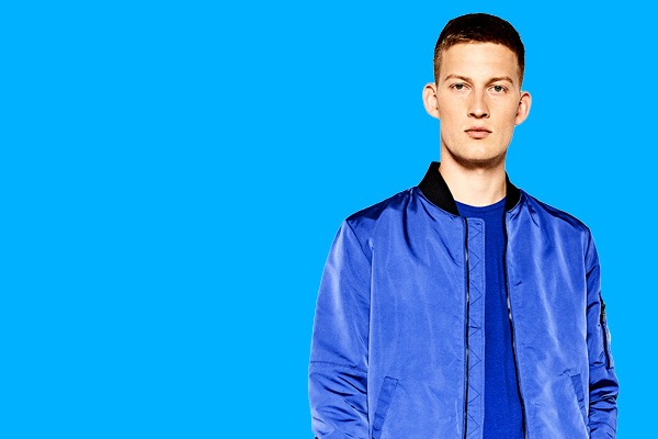 Male model wearing a cobalt blue bomber jacket over a navy crewneck t-shirt, front view against bright blue background